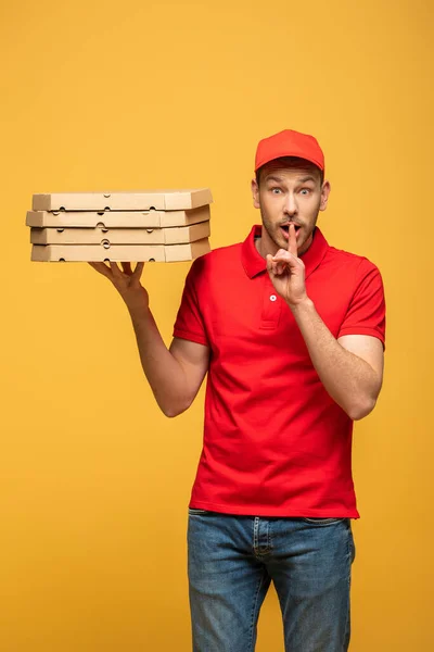 shocked delivery man in red uniform holding pizza boxes and showing shh sign isolated on yellow