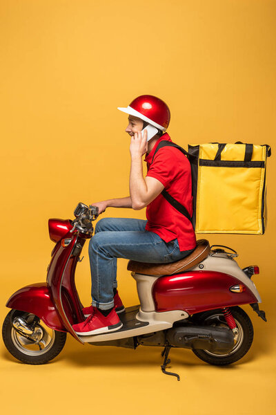 side view of delivery man with backpack on scooter talking on smartphone on yellow background