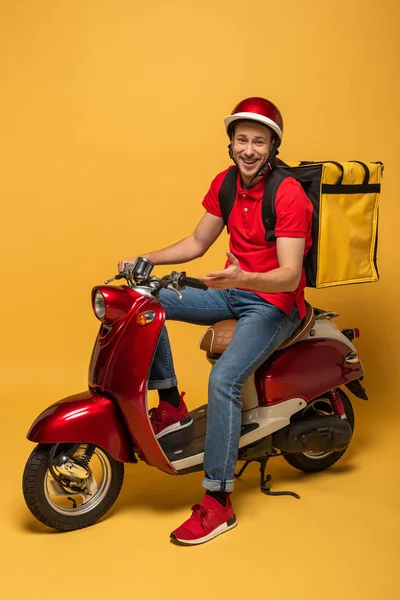 Sarı arka planda motosikletli, sırt çantalı mutlu teslimatçı.