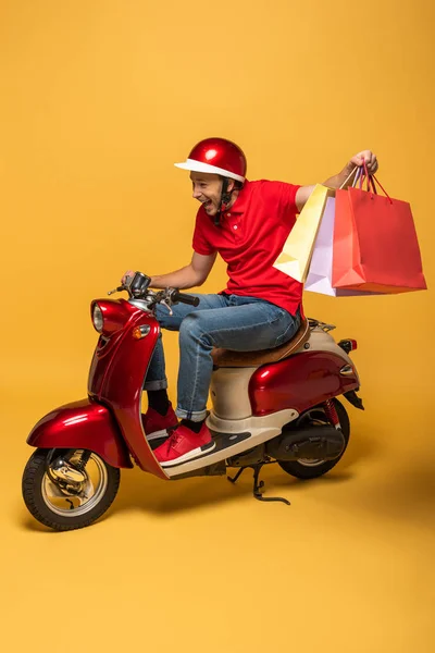 幸せな配達人で赤い制服を着たショッピングバッグでスクーター黄色の背景 — ストック写真