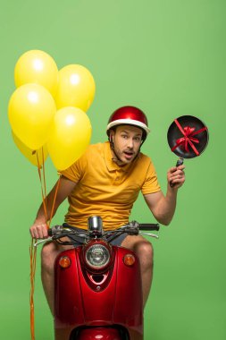 Sarı üniformalı şaşkın teslimatçı motosikletli, tavalı ve yeşil balonlu.