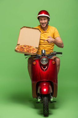 Sarı üniformalı gülümseyen teslimatçı scooter ile yeşil üzerinde pizzayı işaret ediyor.
