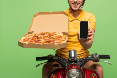 Sarı üniformalı gülümseyen teslimatçının motosikletli ve yeşil üzerinde izole edilmiş akıllı telefonlu görüntüsü.