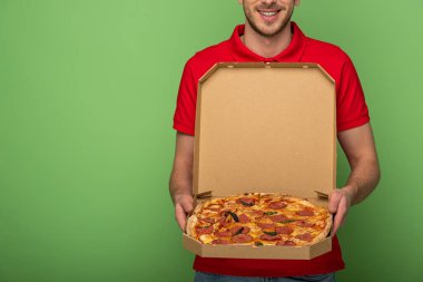 Kırmızı üniformalı gülümseyen teslimatçının yeşil üzerinde pizza kutusu tutarken çekilmiş görüntüsü.