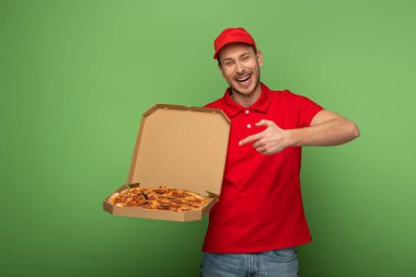 Kırmızı üniformalı mutlu teslimatçı yeşildeki pizza kutusunu işaret ediyor.