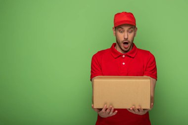 Kırmızı üniformalı teslimatçı yeşilde paket tutuyor.