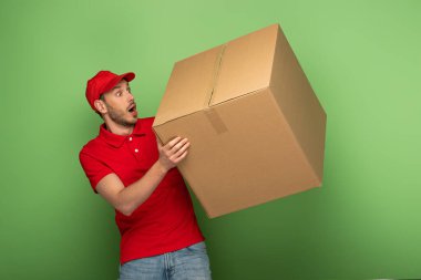 shocked delivery man in red uniform holding huge parcel on green