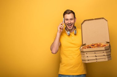 Sarı kulaklıklı mutlu adam pizza tutuyor ve sarı arka planda akıllı telefondan konuşuyor.