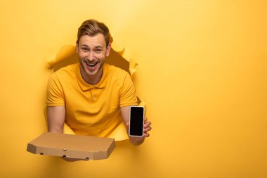 Sarı kıyafetli mutlu yakışıklı adam elinde akıllı telefon ve sarı kağıt delikte pizza kutusu tutuyor. 