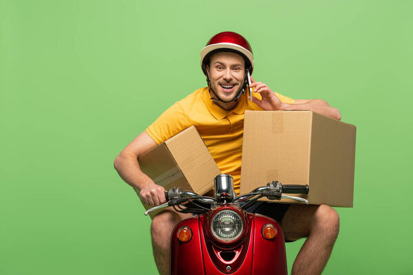 happy delivery man in yellow uniform on scooter with boxes talking on smartphone isolated on green