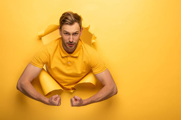 сильный красивый мужчина в жёлтом костюме показывает бицепсы в жёлтом бумажном отверстии
 