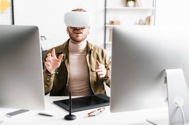 Excited 3d artist in virtual reality headset gesturing near computers and graphics tablet on table clipart