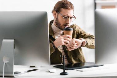 Selective focus of 3d artist looking at wristwatch while drinking coffee near digital devices on table clipart