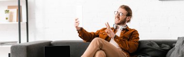 Gülümseyen 3D ressamın panoramik görüntüsü barış işareti gösterirken dijital tablette kanepenin yanındaki bilgisayardan video çağrısı alıyor. 