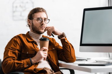 Thoughtful digital designer holding paper cup and stylus near computer monitor on table  clipart