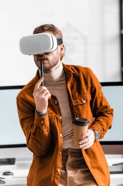 Digital designer in vr headset holding stylus of graphics tablet and coffee to go near computer monitors in office  clipart