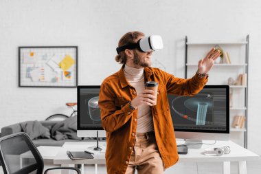 Sanal gerçeklik kulaklığıyla neşeli dijital tasarımcı masadaki bilgisayar monitörlerinde 3D tasarım projesinin yanına gitmek için kahve tutuyor.