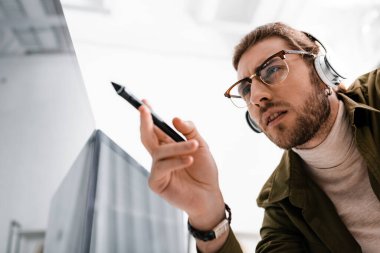 Low angle view of 3d artist in headphones holding stylus of graphics tablet near computer monitor with black screen in office clipart
