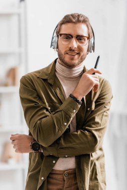 Handsome 3d artist in headphones holding stylus of graphics tablet and smiling at camera in office clipart