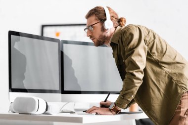 Ofisteki masada VR kulaklığı yanında bilgisayar ve grafik tabletleriyle çalışan 3D kulaklıklı sanatçının yan görüntüsü 