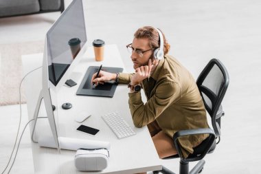 Masada grafik tableti ve bilgisayarlarla çalışırken kulaklık takan 3D sanatçının yan görüntüsü müzik dinliyor