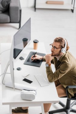 Sanal gerçeklik kulaklığının yanında bilgisayar ve grafik tabletleriyle çalışan kulaklıklı dijital tasarımcının yüksek açılı görüntüsü 