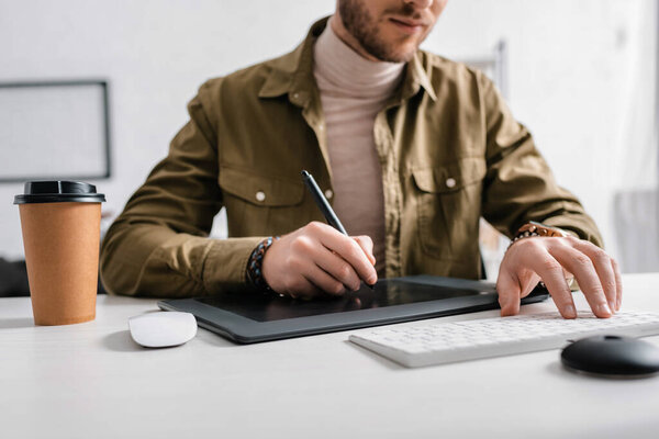 Cropped view of 3d artist using graphics tablet and computer keyboard near coffee to go on table 