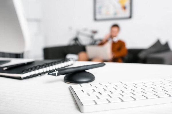Selective focus of computer keyboard, notebook and stylus of graphics tablet on table and designer with laptop on couch in office 