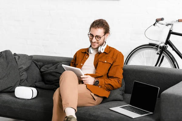 Smiling digital artist using tablet near vr headset and laptop on couch