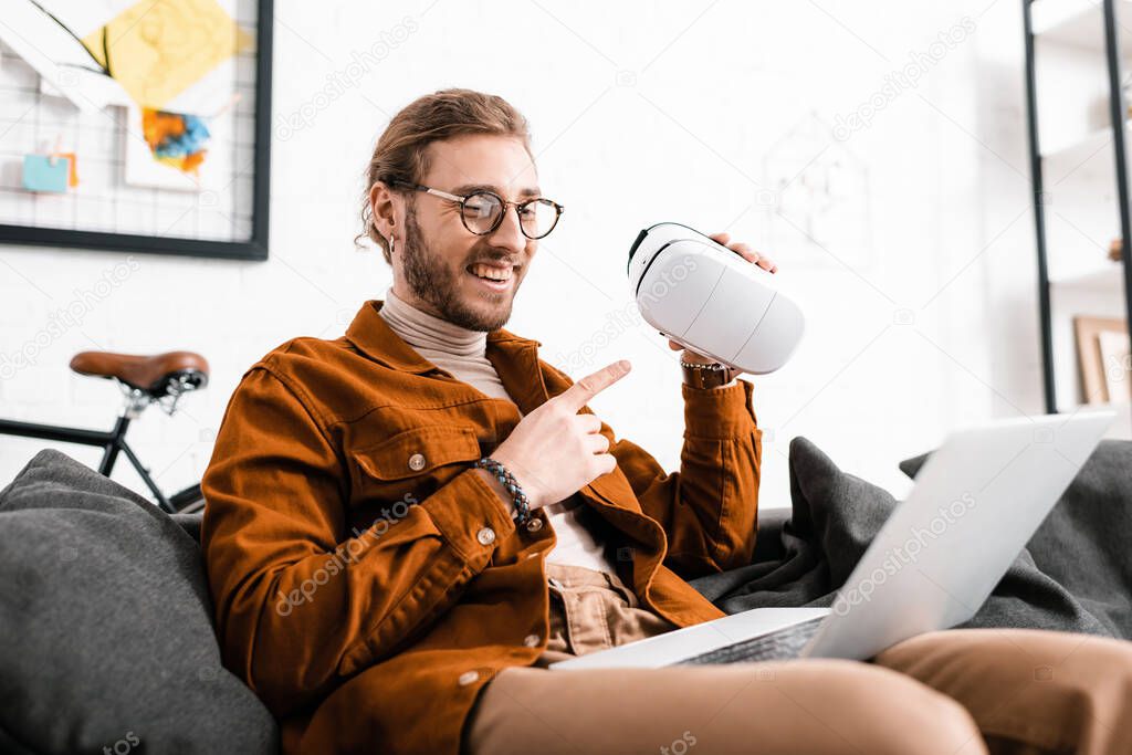 Cheerful 3d artist pointing on virtual reality headset while having video call on laptop on couch 