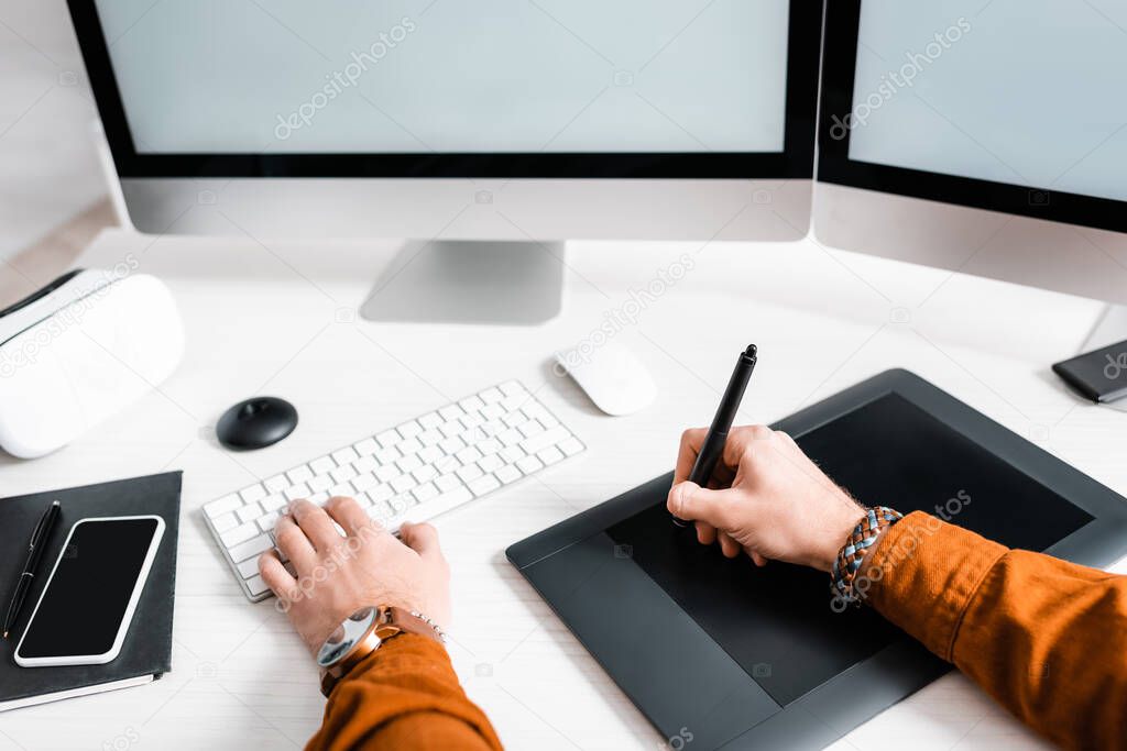 Cropped view of digital designer using graphics tablet near computer monitors, smartphone and vr headset on table