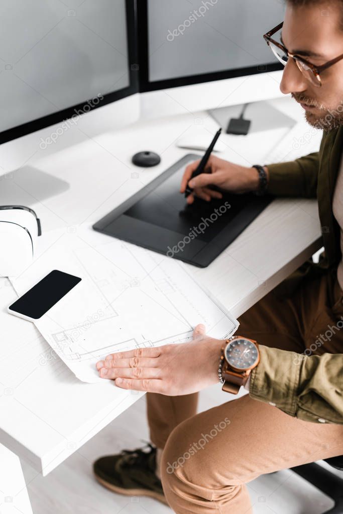 Handsome digital designer using blueprint while working with graphics tablet and computers near vr headset on table