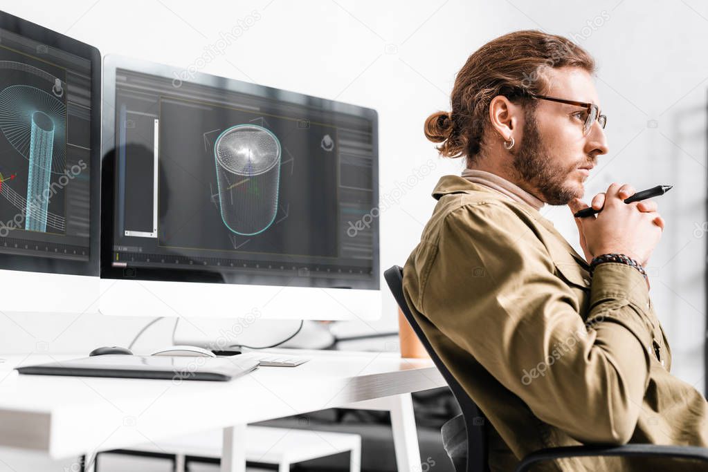 Side view of 3d artist holding stylus of graphics tablet and looking away near project of 3d design on computer monitors on table 