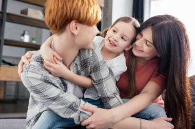 Cheerful same sex couple embracing daughter at home  clipart