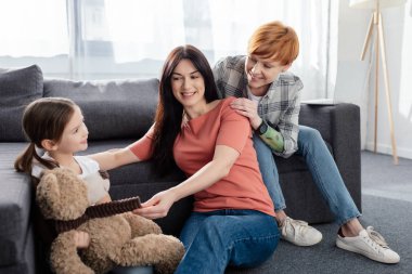 Smiling same sex family sitting near daughter with teddy bear in living room clipart