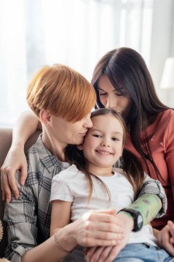 Same sex parents kissing daughter smiling at camera on couch at home clipart
