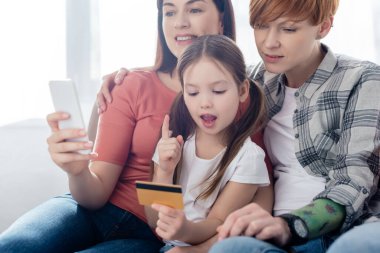 Evdeki kanepede kredi kartıyla, akıllı telefonlu, aynı cinsiyetten ailelerin gülümsemesi. 