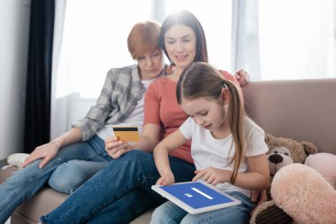 Kyiv, Ukrayna - 10 Şubat 2020: Çocuk facebook uygulamalı dijital tablet kullanıyor kanepesinde kredi kartı olan annelerin yanında.
