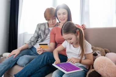 Kyiv, Ukrayna - 10 Şubat 2020: Instagram uygulamalı dijital tablet kullanan çocuk, yanında kanepesinde kredi kartıyla gülümseyen annelerin olduğu ekranda 