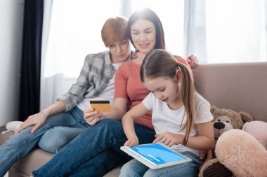 KYIV, UKRAINE - FEBRUARY 10, 2020: Child using digital tablet with twitter app on screen near smiling same sex parents with credit card on sofa