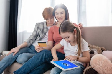 KYIV, UKRAINE - FEBRUARY 10, 2020: Kid using digital tablet with shazam app on screen near same sex parents with credit card on sofa at home