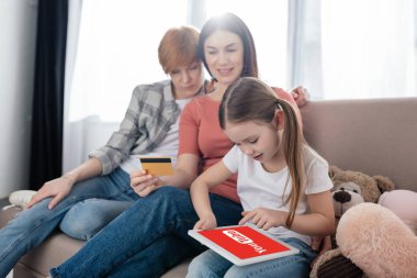 KYIV, UKRAINE - FEBRUARY 10, 2020: Child using digital tablet with youtube app on screen near moms with credit card on couch