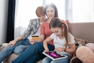 Kronometre uygulamalı dijital tablet kullanan bir çocuk annesinin yanında oturma odasındaki koltukta kredi kartıyla oturuyor.
