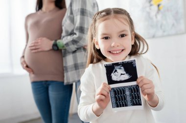 Selective focus of smiling kid holding ultrasound scan of baby near mom embracing pregnant woman at home  clipart