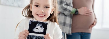 Selective focus of smiling kid holding ultrasound scan of baby near mother embracing pregnant woman at home, panoramic shot clipart