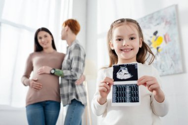 Selective focus of smiling kid holding ultrasound scan of baby near woman embracing pregnant girlfriend  clipart