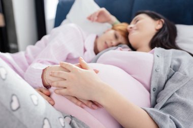 Selective focus of woman touching belly of pregnant girlfriend while sleeping on bed  clipart