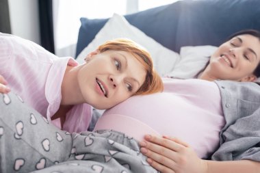 Selective focus of woman listening belly of smiling pregnant girlfriend on bed in bedroom clipart