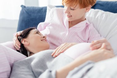 Selective focus of woman smiling at pregnant girlfriend in pajamas on bed at home clipart