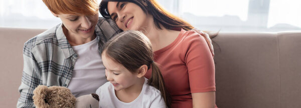 Panoramic shot of smiling same sex couple sitting near daughter with teddy bear on couch